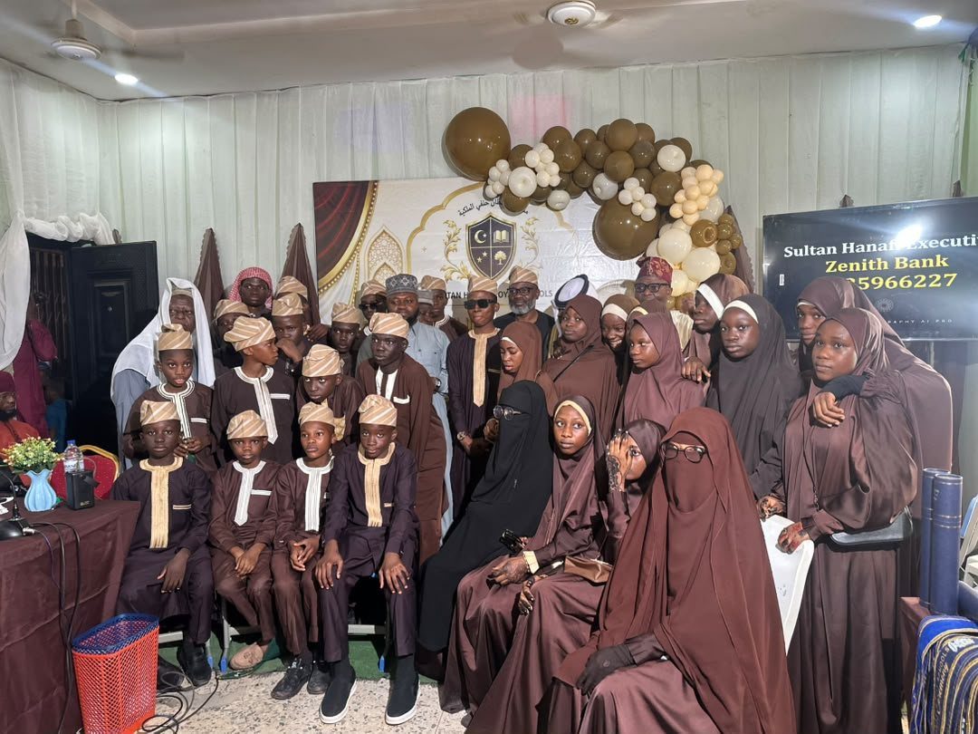 The graduating Hifz cohort of Sultan Hanafi Qur'an College, boys in brown and cream and girls in brown, gathered before the school's banner at the graduation ceremony